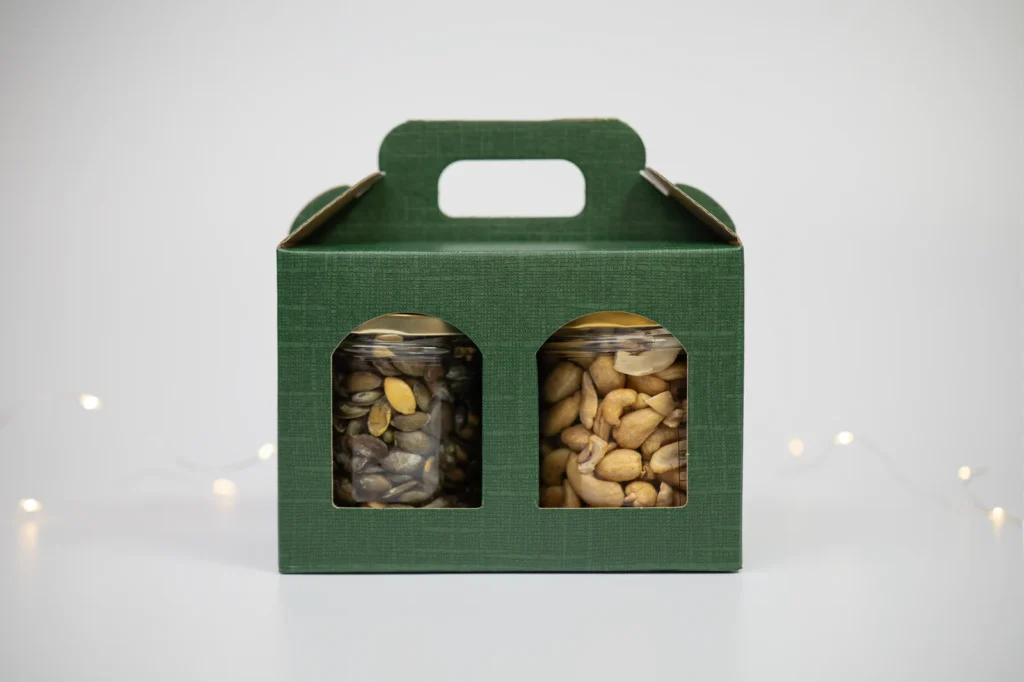 Green textured gift box with a built-in handle, two front windows showing jars of mixed nuts inside; festive backdrop with soft string lights.